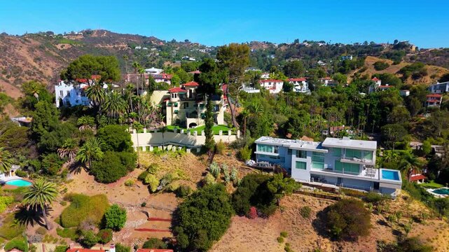 Aerial drone footage pans right to left over luxury hillside homes in Hollywood Hills, revealing a Spanish colonial mansion with terracotta roof tiles beside a modern glass house with a swimming pool