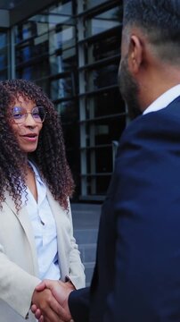 Vertical. Black Latina professional shaking hands with a male colleague outside a modern office while coworkers gather around. Corporate scene conveying networking diverse professional collaboration