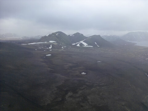 Aerial view of the volcanic landscape featuring dark terrain, patches of snow, and distant mountains under a cloudy sky, Iceland.