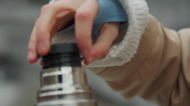 Close up of hand wearing fingerless gloves gripping lid of stainless steel thermos. person is dressed in warm clothing suggesting outdoor setting with soft lighting and muted background