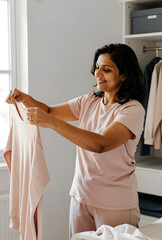 Obraz premium Home organization and daily routine, smiling indian woman folding laundry in a bright bedroom closet