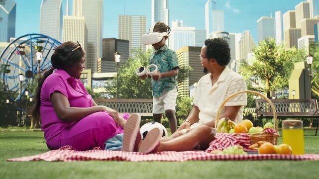 Small boy explores virtual reality with a VR headset and handheld controllers while parents watch from a picnic blanket in a sunny urban park, combining immersive and interactive technology outdoors.