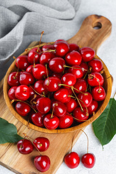 Fresh ripe sweet cherries in wooden bowl on rustic cutting board with green leaves and linen cloth on white textured background. Healthy summer fruit, red berries, fresh seasonal harvest, healthy food