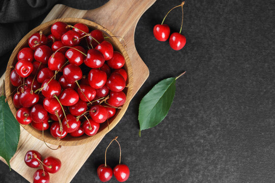 Fresh ripe sweet cherries in rustic wooden bowl with green leaves on black textured background. Top view flat lay with copy space. Juicy organic summer fruit, red berries, healthy seasonal food
