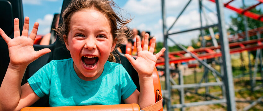 Excited girl riding roller coaster with hands up screaming with joy. Happy kid enjoying thrilling amusement park attraction. Childhood adventure and summer vacation entertainment concept.