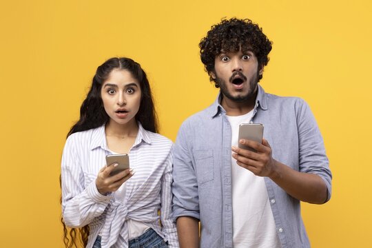 Surprised Expressions From Young Couple, Male And Female, In Their 20s Or 30s, Holding Smartphones And Looking At Screens With Shocked Faces, In A Studio Setting. Digital Communication Concept.