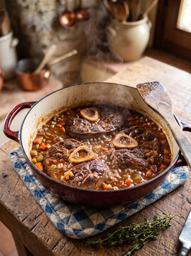 Hot bubbling osso buco in red cast iron pot on rustic wooden kitchen table with rising steam