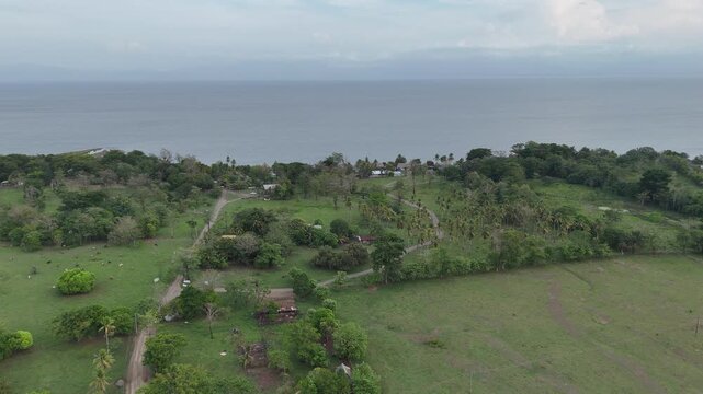 Drone flies forward over green pastures and a dirt road toward Lake Izabal surrounded by lush tropical vegetation in El Estor, Guatemala.