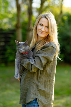 Blonde woman holds a Russian Blue cat outdoors in a green garden. Relaxed portrait of a pet owner with her gray pedigree cat in soft natural sunlight.