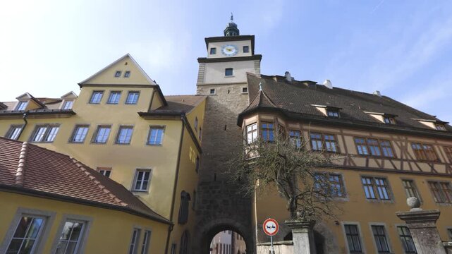 4K footage of a historic clock tower surrounded by traditional medieval buildings in Rothenburg ob der Tauber, Bavaria, Germany, under a clear blue sky.
