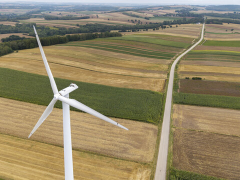 Aerial view of a white wind turbine standing amidst a patchwork of agricultural fields and a rural road in Poland.