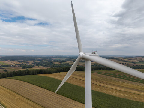Aerial view of a wind turbine standing amidst vast agricultural fields and rolling hills under a cloudy sky in Poland.