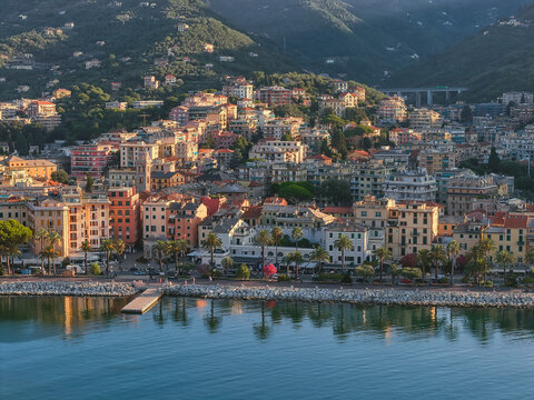Aerial view of the coastal town of Rapallo with colorful Mediterranean buildings, palm trees along the waterfront, and rocky breakwater under golden hour light in Rapallo, Liguria, Italy.