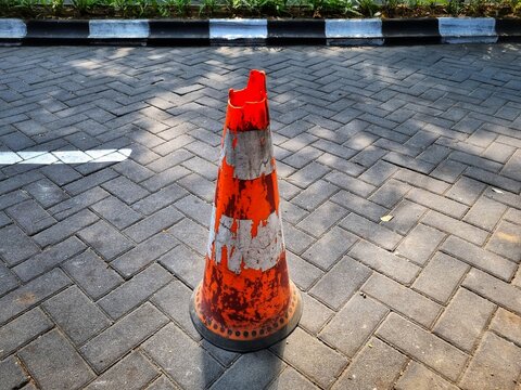 Weathered orange traffic cone with peeling stripes and broken at the tip, on grey paving block road.
