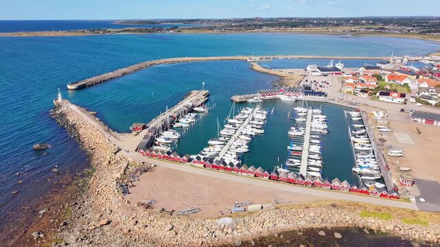 Aerial view of Traslovslage harbor with rows of red fishing huts, docked sailboats, and a stone breakwater along the coastline in Traslovslage, Halland County, Sweden.