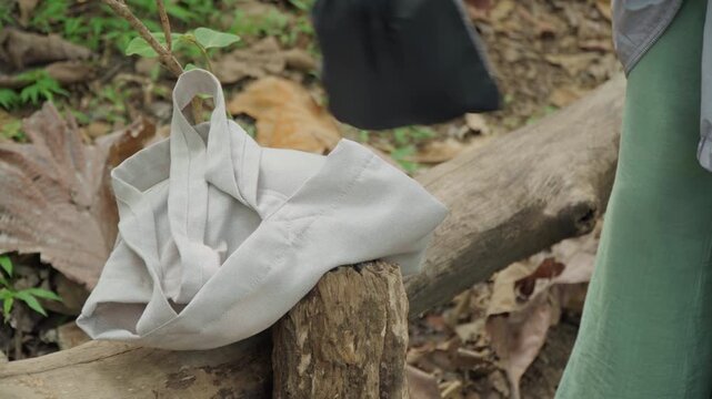 Close up of person placing bags on wooden logs in outdoor setting. white bag and drawstring pouch are arranged near tree stumps with green pants visible. Background includes dry leaves soil