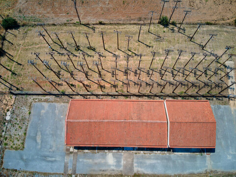 Aerial view of an electrical substation with rows of utility poles casting long shadows, a red-tiled roof building, and gravel ground in Kassandreia, Greece.