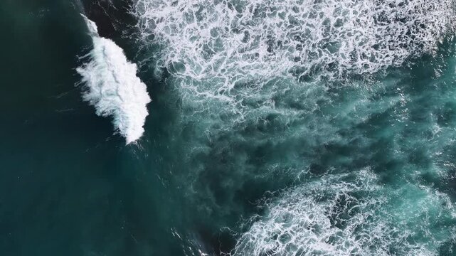 Aerial view of turquoise ocean waves crashing and creating white sea foam against the deep blue water in Kuta Selatan, Bali, Indonesia.