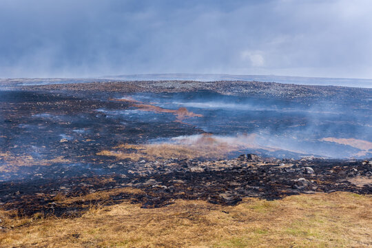 Smoldering ground and burnt vegetation following wild hill fire, light smoke drifting over charred landscape under dramatic overcast sky