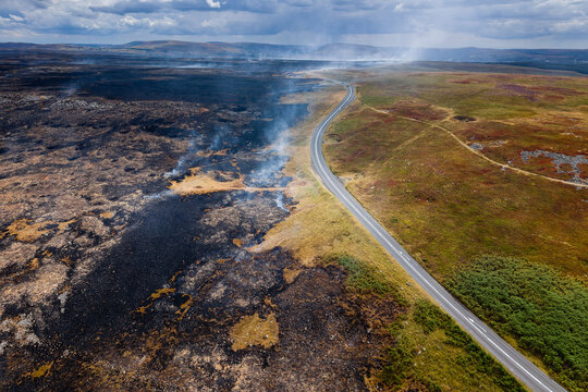 Blackened earth and smoldering grass following wild moorland fire, sharp divide between burnt area and green terrain along country highway