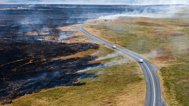 Wildfire aftermath along rural road, charred scorched earth and smoke rising beside untouched grassy moorland, high angle aerial view