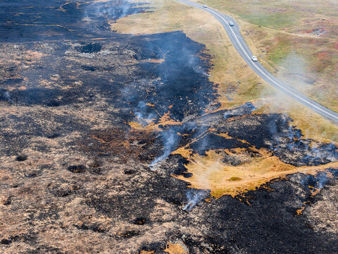 Charred moorland ground smoldering after grass fire, dark burnt patches contrasting with dry yellow vegetation along upland pass road