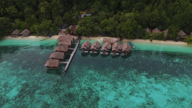 Aerial view of Mios Kri overwater bungalows with thatched roofs situated over clear turquoise water along a lush tropical coastline in West Supiori, Papua, Indonesia.