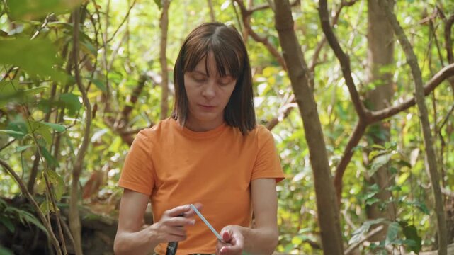 Close up of woman in orange shirt handling water bottle and small blue object amidst dense green foliage. Woman manipulates items with steady hands surrounded by natural forest environment