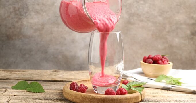 Woman pouring tasty raspberry smoothie from jug into glass at wooden table, closeup