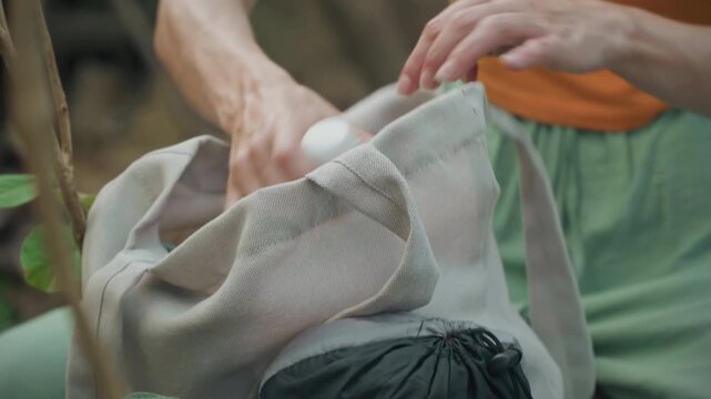 Close up of woman in orange top and green pants unpacking items from fabric bag placing water bottle inside beige sack. Hands interact with plastic wrapped bread while adjusting gray