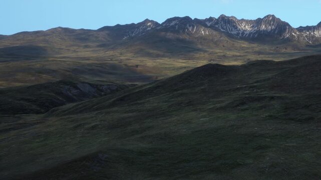 Green hills stretch under a clear blue sky. Distant mountains loom with snow on top. The scene is quiet with no people around. Light changes as the sun sets over the landscape.