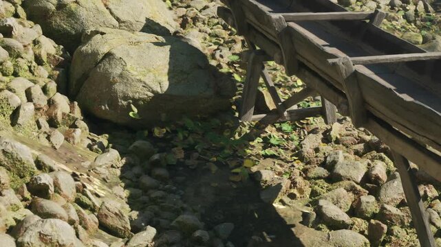 A wooden bridge is positioned above a small stream. The area is filled with rocks and green leaves. Sunlight shines on the water, illuminating the natural surroundings.