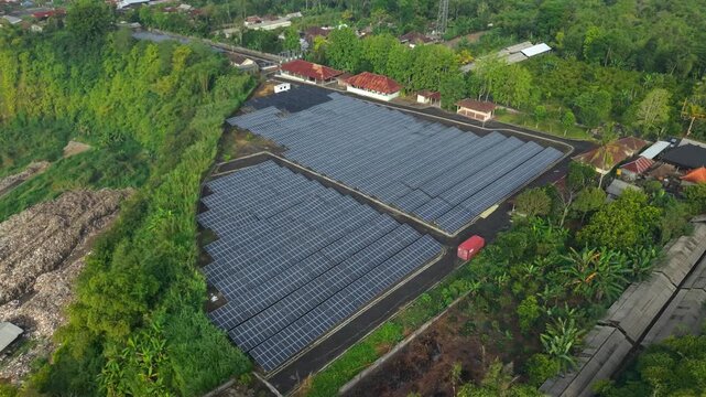 Aerial view of the PLTS 1MWp Bangli solar farm featuring rows of photovoltaic panels surrounded by lush green vegetation and trees in Bangli, Bali, Indonesia.