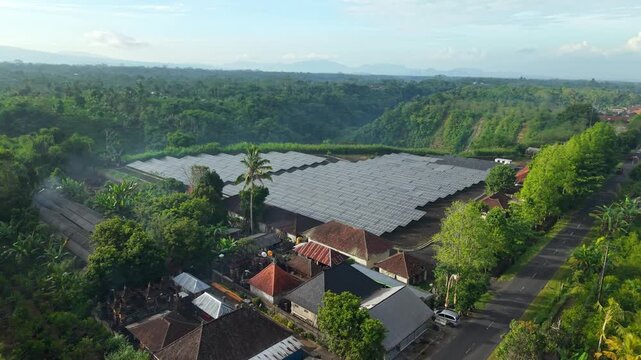 Aerial view of the PLTS 1MWp Bangli solar power plant featuring rows of photovoltaic panels surrounded by lush tropical vegetation and traditional buildings in Bangli, Bali, Indonesia.