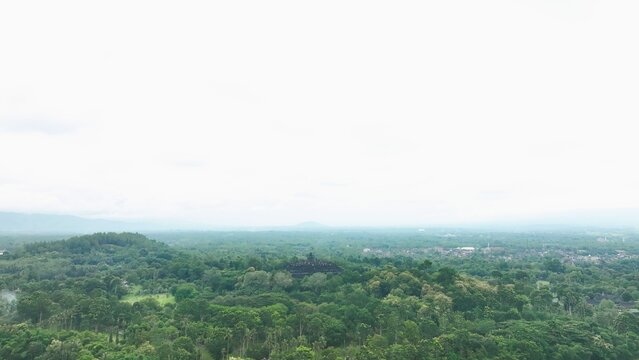 Aerial view of the ancient Borobudur temple complex nestled within a vast expanse of lush green tropical forest under a soft overcast sky in Borobudur, Central Java, Indonesia.