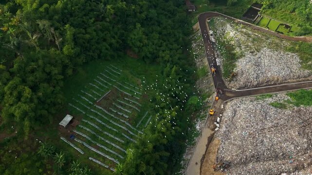 Aerial view of TPA Bangli landfill site with surrounding lush forest, dirt roads, and birds flying over the waste management area in Bangli, Bali, Indonesia.
