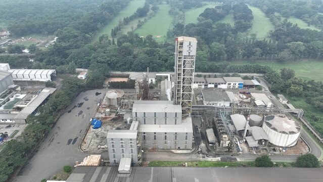 Aerial view of an industrial manufacturing plant featuring a tall processing tower, storage tanks, and surrounding green landscape in Cikampek, West Java, Indonesia.