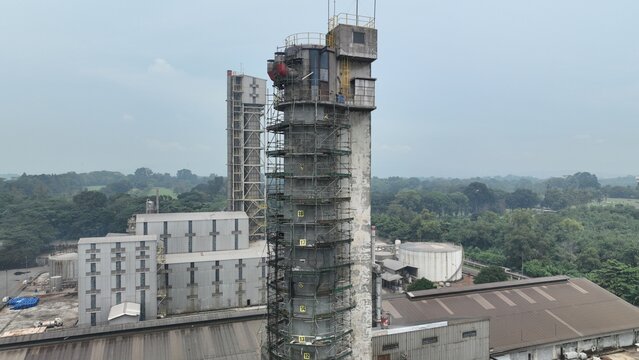 Aerial view of an industrial facility featuring a tall tower covered in scaffolding for maintenance, surrounded by factory buildings and lush green trees in Cikampek, West Java, Indonesia.