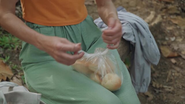 Close up of woman in orange top and green pants opening plastic bags with bread while sitting outdoors on ground. gray jacket rests nearby indicating travel or outdoor activity. scene is set