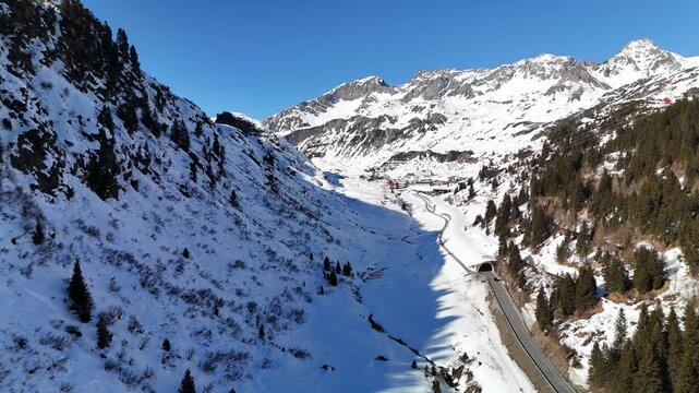 Aerial view of Arlberg mountain pass with snow-covered slopes, pine forests, and a road leading into a tunnel under a clear blue sky in Bludenz District, Vorarlberg, Austria.
