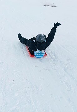 Happy person riding red snow sled toboggan down snowy hill with arm raised in winter fun