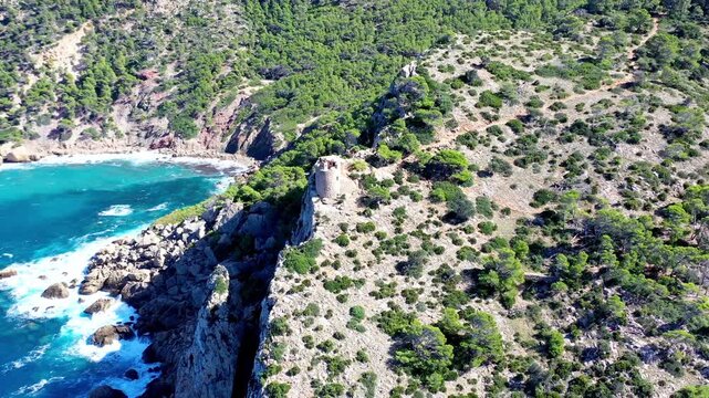 Aerial drone view over rocky coastline of Mallorca featuring ancient watchtower above turquoise Mediterranean sea waters.