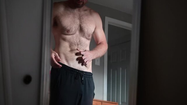 Man after weight lose looking at his reflection in the mirror, muscular with loose skin, dad bod, body transformation, stretch marks belly lose skin