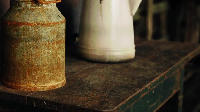 A rusty milk can sits next to a white kettle on a wooden shelf. The scene shows a simple display of old kitchen items in a rural setting. The lighting adds warmth to the display.