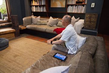 Fototapeta premium Senior Asian man sitting on suede sofa in living room using tablet near bookshelves