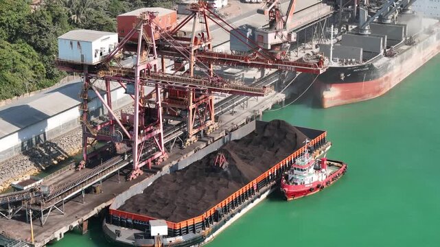 Aerial view of a coal loading terminal with a barge being loaded by a conveyor system and a tugboat moored alongside in Indonesia.