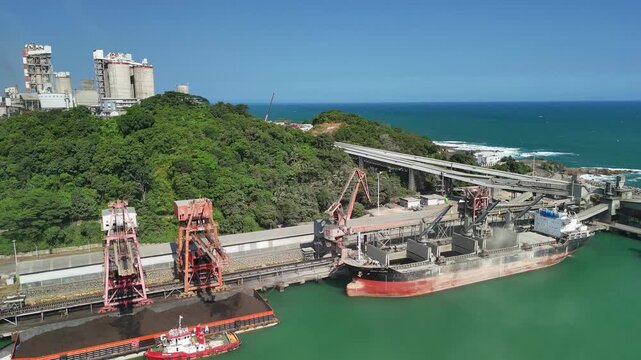 Aerial view of a large cargo ship being loaded with bulk materials by industrial cranes at a coastal port facility in Indonesia.