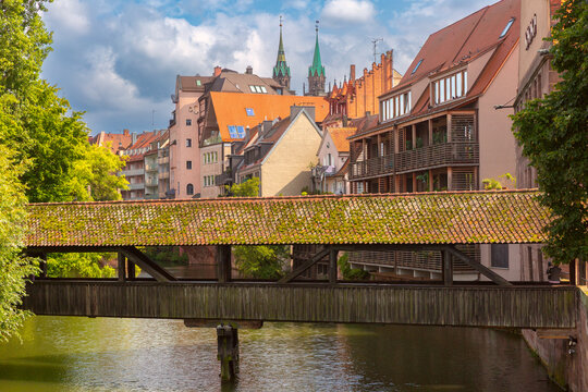 Traditional covered wooden bridge crosses the Pegnitz River among historic buildings and green trees in Nuremberg, Germany.