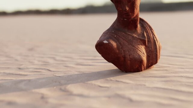 A small sculpture is placed on a sandy surface as the sun sets in the background. The light creates long shadows and highlights the texture and shape of the sculpture.