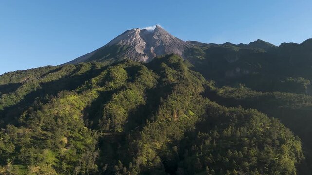 Aerial view of Mount Merapi volcano with lush green forested slopes and rocky peak under a clear blue sky in Cangkringan, Special Region of Yogyakarta, Indonesia.